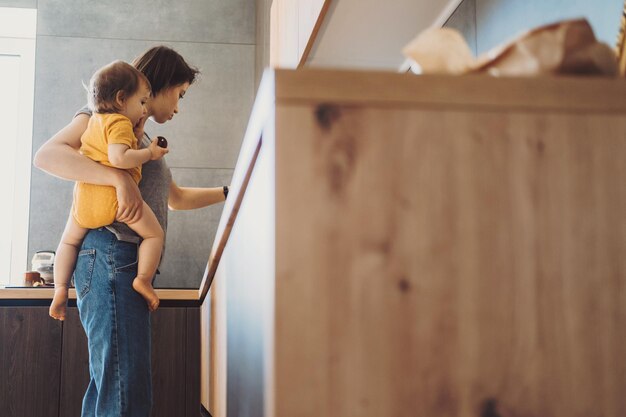 Side view of a mum holding her young baby girl while she making food in her kitchen. Family care. Family protection. Baby care.