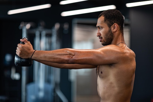 Вид сбоку мускулистого чернокожего мужчины без рубашки, тренирующегося с гирями в спортзале