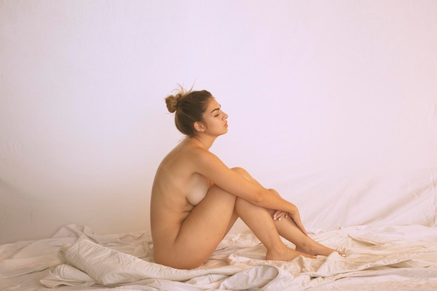 白い壁に逆らってベッドに座っている裸の若い女性のサイドビュー