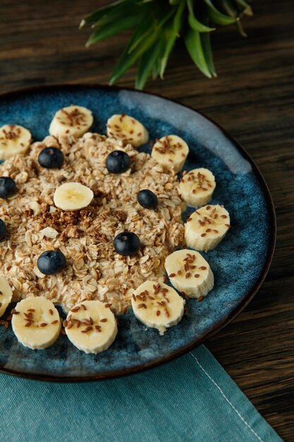 Side view of oatmeal with banana blackthorn flax and sesame in plate on blue cloth with pineapple leaves on wooden background