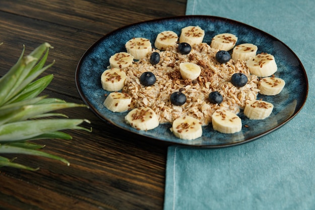 Side view of oatmeal with banana blackthorn walnut and sesame in plate on blue cloth with pineapple leaves on wooden background