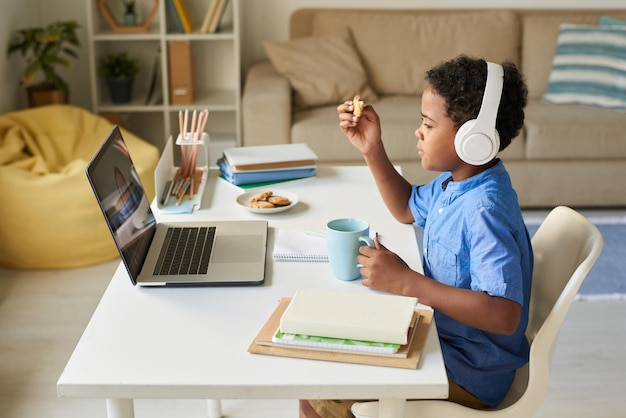Фото Боковой вид афроамериканского мальчика в беспроводных наушниках, сидящего за домашним столом и едящего печенье во время разговора с онлайн-учителем