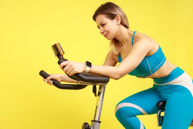 Фото Вид сбоку на спортивную женщину, катающуюся на велотренажере и смотрящую обучающее видео на кардио-тренировке смартфона в синей спортивной одежде. снимок в студии на желтом фоне
