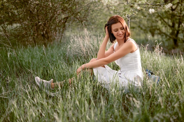 Фото Брюнетка в белом платье сидит на траве с закрытыми глазами и трогает волосы на лугу летом, вид сбоку