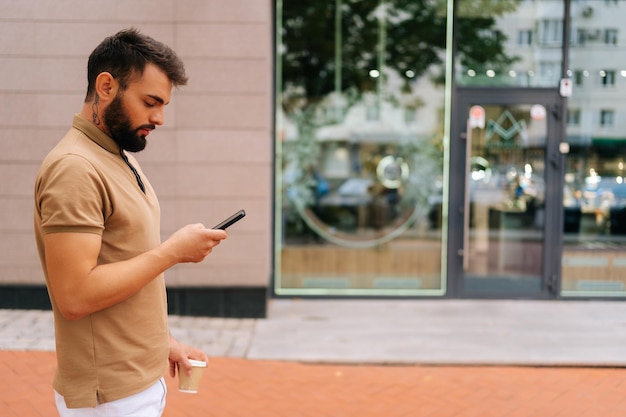 Фото Вид сбоку уверенного в себе молодого человека в повседневной одежде, идущего по городской улице с помощью мобильного телефона и пьющего кофе на вынос в летний день