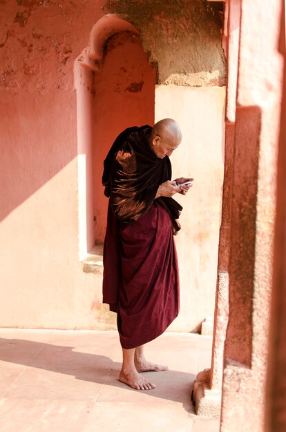 Фото Боковой вид монаха, использующего смартфон, стоящего у стены