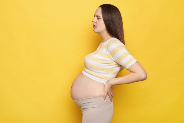 写真 病気の疲れた白人の妊婦の横の景色 裸の腹でカジュアルなトップを着て 黄色の背景に隔離され 背中を触って 不快感と疲労を感じています