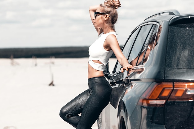 Фото Вид сбоку на тощую грациозную женщину в черных брюках и солнцезащитных очках рядом с черной машиной, смотрящую вперед