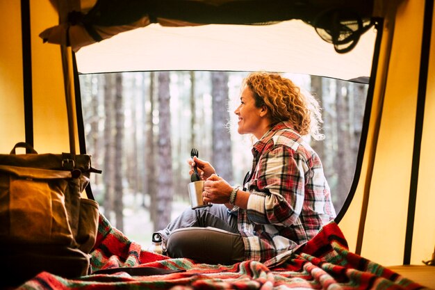 Фото Боковой вид улыбающейся женщины с кружкой в руках, сидящей у палатки в лесу