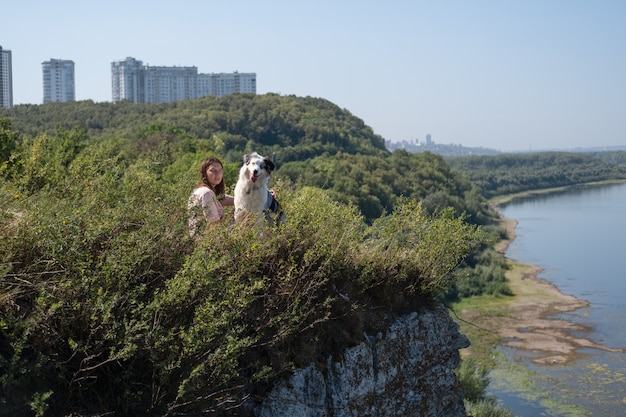 사진 여름 강둑에 오스트레일리아 셰퍼드 블루 멀 개와 함께 앉아 있는 여성의 모습. 인간과 동물 사이의 사랑과 우정. 애완 동물과 함께 여행하십시오.