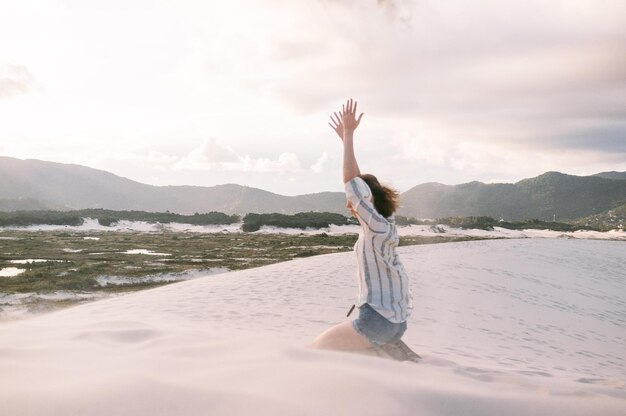 写真 雲の空を背景に砂の上にひざまずいて腕を挙げた女性の横の景色