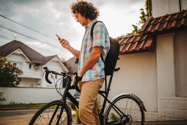 巻き毛の若い幸せな男性の側面図彼の携帯電話で自転車の道路のgpsアプリケーションを選択スマートフォンを使用してバックパックサーフィンインターネットで幸せな笑顔の男のサイクリスト