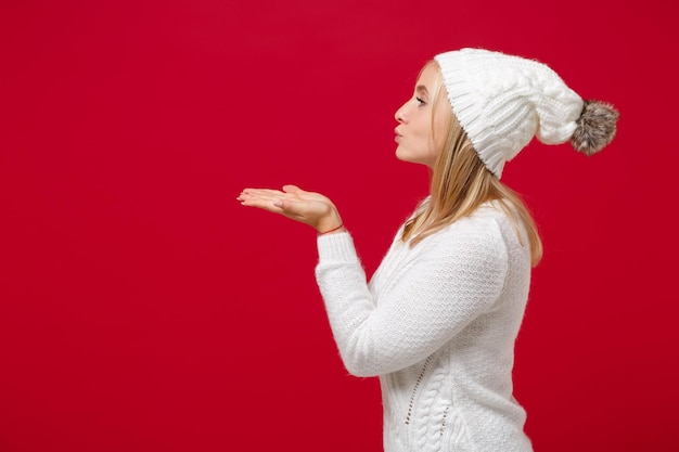 사진 스튜디오의 빨간색 배경에 격리된 흰색 스웨터 모자를 쓴 젊은 여성의 측면. 건강한 패션 라이프 스타일 추운 계절 개념. 복사 공간을 비웃습니다. 공기 키스를 날리며 손에 무언가를 잡습니다.