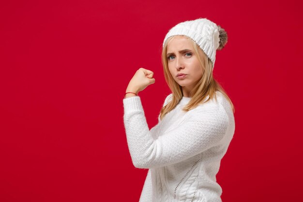 写真 白いセーターの若い女性の側面図、赤い背景のスタジオ ポートレートに分離された帽子。健康的なファッション ライフ スタイル、人々 の感情、寒い季節のコンセプト。コピー スペースをモックアップします。上腕二頭筋、筋肉を示します。