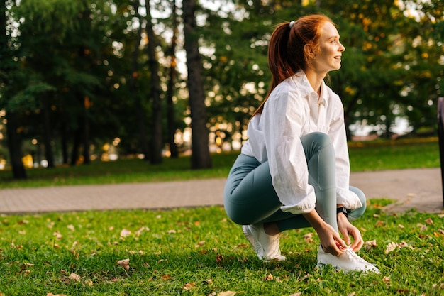 Side view og cheerful redhaired young woman tying shoelaces on sneakers before running