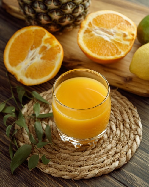 Side view of orange juice in glass with leaves on trivet and half cut oranges with lemon pineapple on cutting board on wooden background