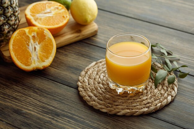 Side view of orange juice in glass with leaves on trivet and half cut oranges with lemon pineapple on cutting board on wooden background