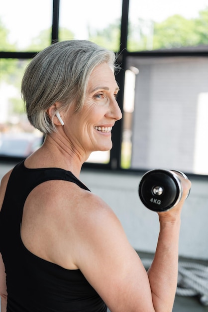 side view of overjoyed senior sportswoman in wireless earphone working out with dumbbell in gym
