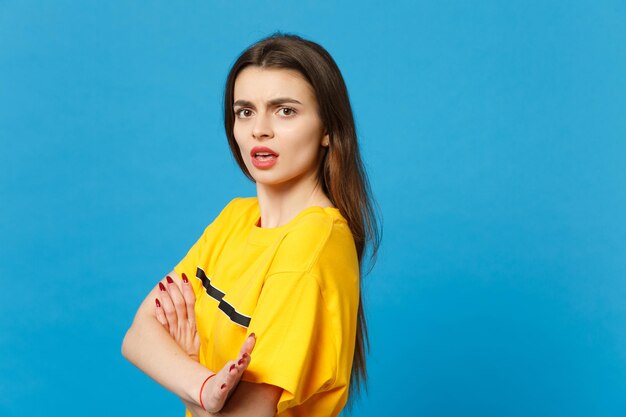 Side view of perplexed irritated young woman in vivid casual clothes looking camera, holding hands crossed isolated on bright blue background in studio. People lifestyle concept. Mock up copy space.