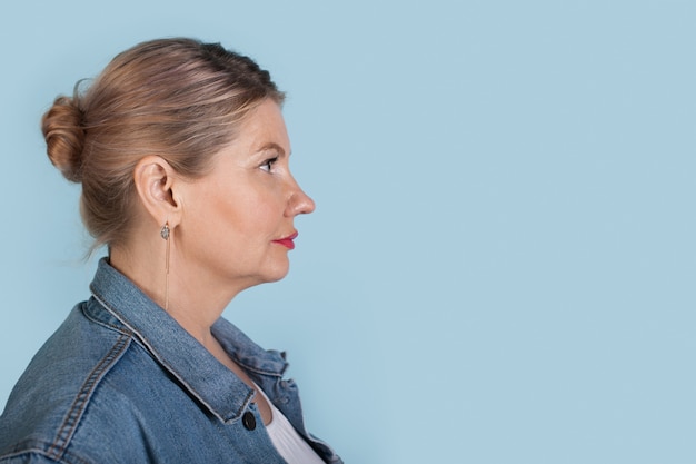 La foto di vista laterale di una donna caucasica senior in cappotto di jeans sta pubblicizzando qualcosa su una parete blu con spazio libero