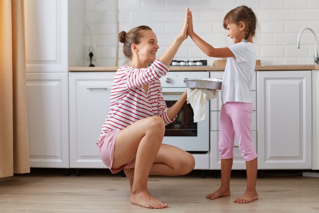 Side view photo of cheerful charming mommy and small kid giving\
high-five in light kitchen, baking delicious dessert together,\
family wearing casual style clothing, posing at home.