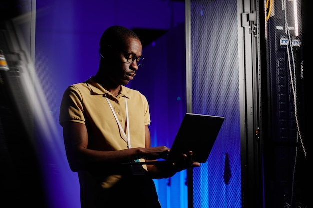 Side view portrait of adult african american man using laptop in dark server room lit by neon light