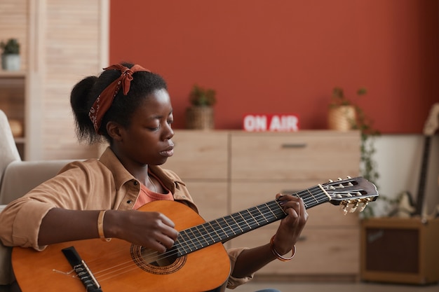 Side view portrait of African-American woman playing acoustic guitar while sitting on floor at home, copy space