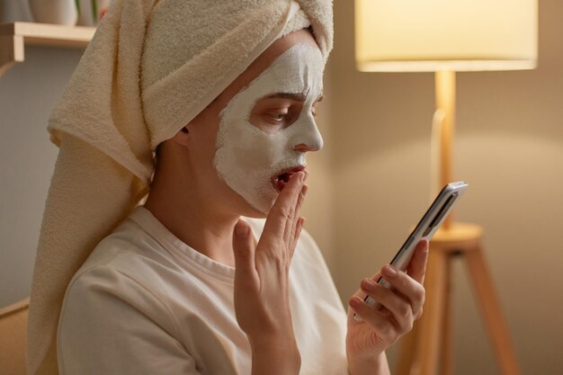 Side view portrait of amazed Caucasian woman with peeling mask on face in white towel sitting on sofa at home doing cosmetic procedures holding smart phone looking at display covering mouth with hand