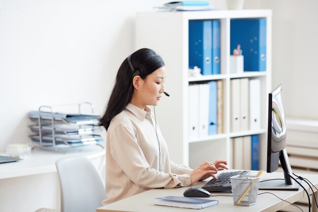 Photo side view portrait of asian woman wearing headset and using computer while working in customer support service hotline