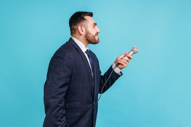 Photo side view portrait of bearded young adult man wearing official style suit standing and offering microphone, asking questions, interviews. indoor studio shot isolated on blue background.