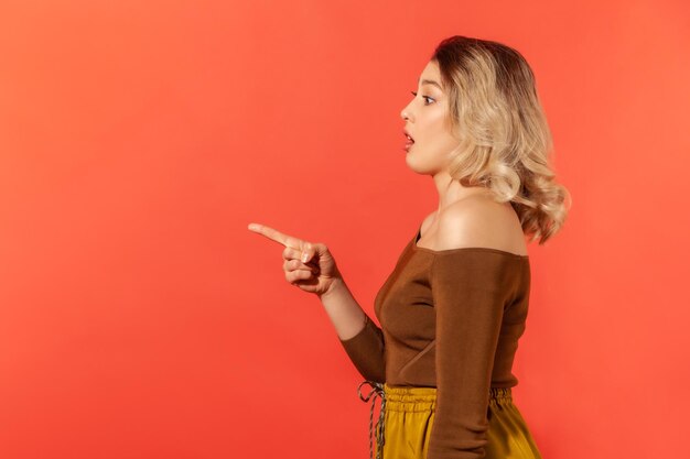Side view portrait of beautiful suprised woman with blonde hair in brown blouse pointing finger to the left and opened mouth looking with astonishment indoor studio shot isolated on red background