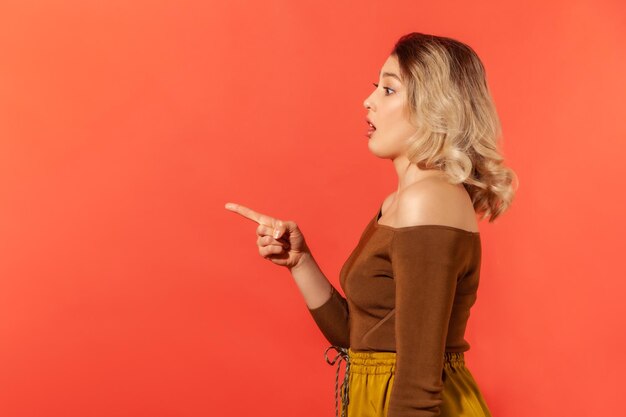 Side view portrait of beautiful suprised woman with blonde hair in brown blouse pointing finger to the left and opened mouth looking with astonishment Indoor studio shot isolated on red background