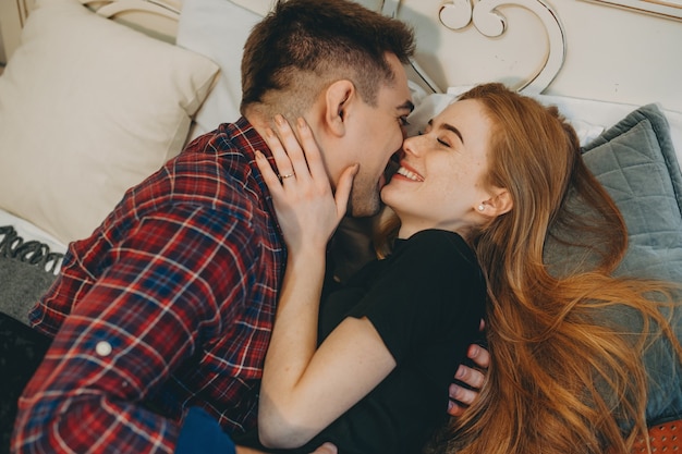 Side view portrait of a beautiful young couple embracing and laughing while leaning on a bed face to face at their home.