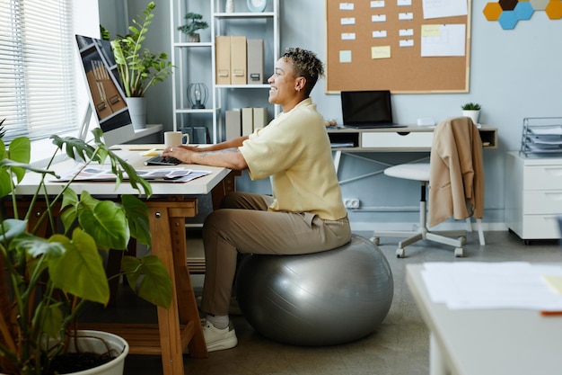 Side view portrait of black young woman sitting on fitness ball at workplace and smiling sports acti