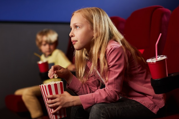 Ritratto di vista laterale dell'adolescente biondo che guarda film nel cinema e che mangia popcorn, spazio della copia