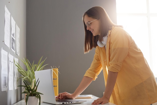 Вид сбоку портрет занятой улыбающейся женщины в желтой рубашке, использующей портативный компьютер и стоящей возле стола в офисе, наслаждающейся завершением работы и выключением ноутбука