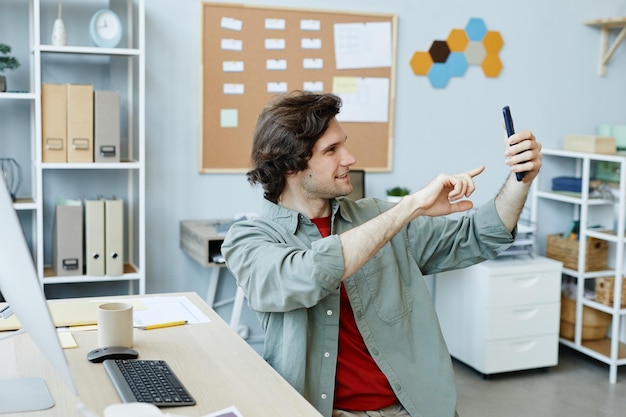Side view portrait of caucasian young man taking selfie photo\
at workplace in office