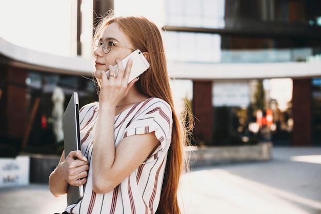 ラップトップを持って建物に対して外のスマートフォンで話している魅力的な若いビジネス女性の側面図の肖像画。