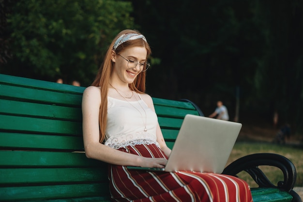 Side view portrait of a charming young red-haired woman looking at a laptop screen laughing while sitting on a bench in the parc.