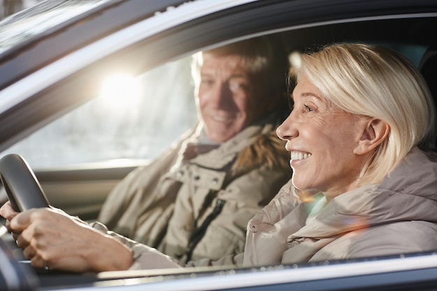 Side view portrait of cheerful senior couple driving car in winter focus on smiling woman behind whe