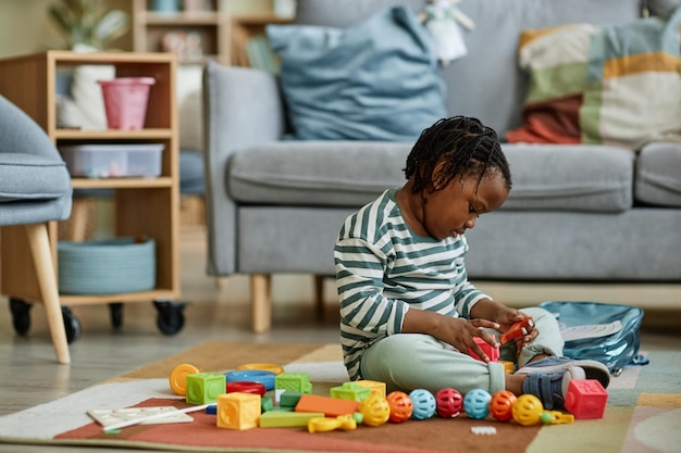 Side view portrait of cute black baby playing with toys while sitting on floor in cozy home copy spa