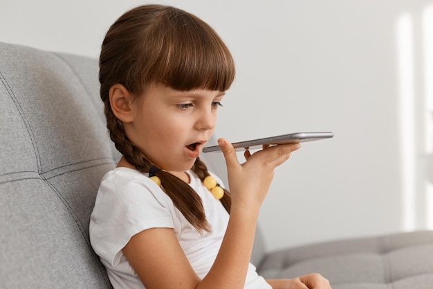 Side view portrait of cute female kid wearing casual style clothing posing in light room and recording message, holding mobile in hands, using voice assistant application.