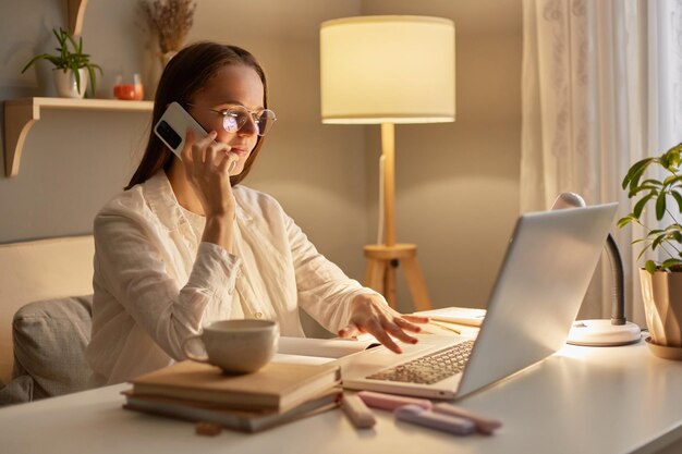 Side view portrait of delighted attractive woman with brown hair sitting at table working on laptop in evening at home and talking with on smart phone having conversation