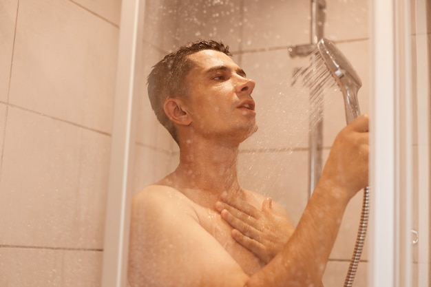 Photo side view portrait of excited man standing under cold shower with open mouth, posing naked in bathroom, refreshing after hard working day, hygiene procedures.