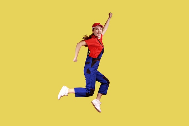 Side view portrait of extremely happy worker woman jumping and clenched fists, celebrating long awaited day off, wearing overalls and red cap. Indoor studio shot isolated on yellow background.