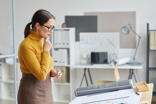 Side view portrait of female architect looking at blueprints while standing by drawing table at workplace, 