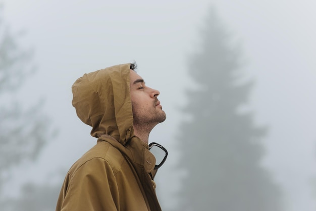 Ritratto di vista laterale del maschio bello che cammina nella natura nebbiosa ascoltando la musica sulle sue cuffie fuori uomo del viaggiatore che indossa il cappuccio del cappotto alla moda in montagna emozioni della gente e concetto di viaggio