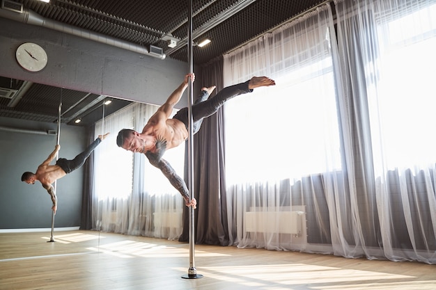 Side view portrait of handsome strong male wearing grey pants doing exercises on the pylon while he is shifting the weight to his arms