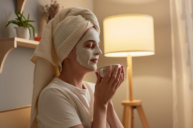 Side view portrait of happy delighted woman with cosmetology mask on face sitting on cough holding cup of coffee and enjoying aromat relaxing at home