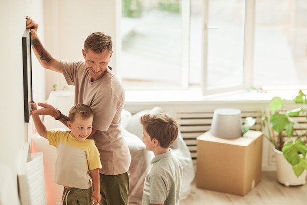 Side view portrait of happy father with two sons hanging pictures on wall while moving in to new hom...